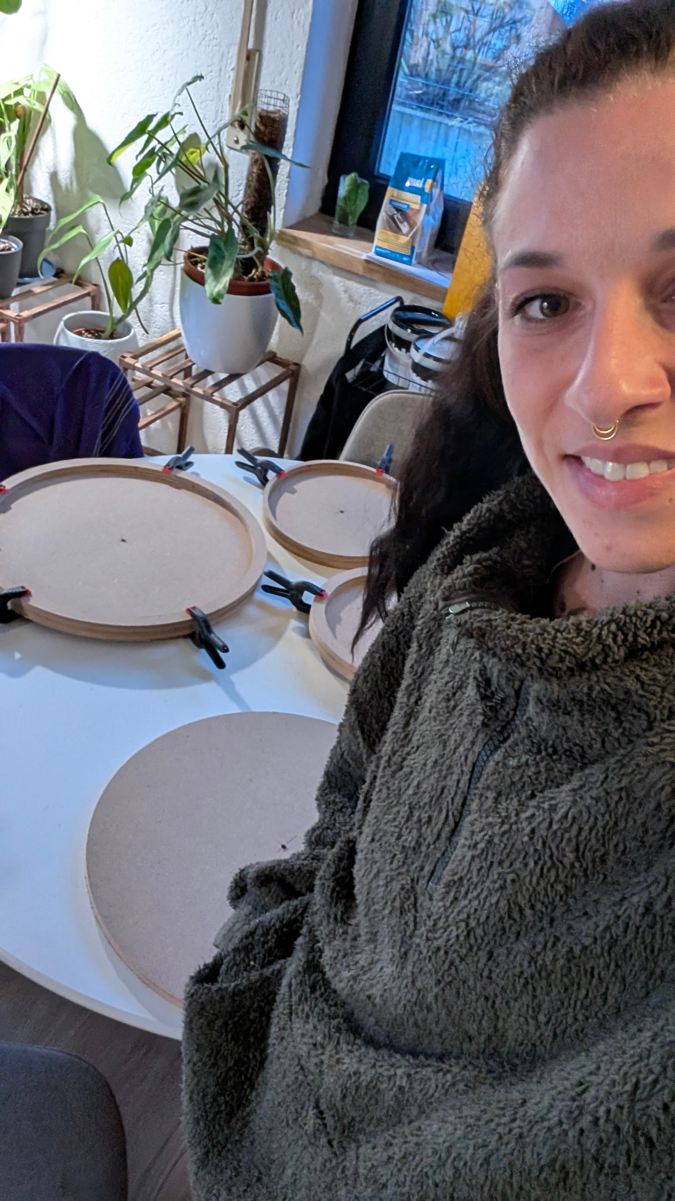 Lächelnde Frau in flauschigem Fleece neben runden Holzbrettern mit Klemmen auf einem weißen Tisch.