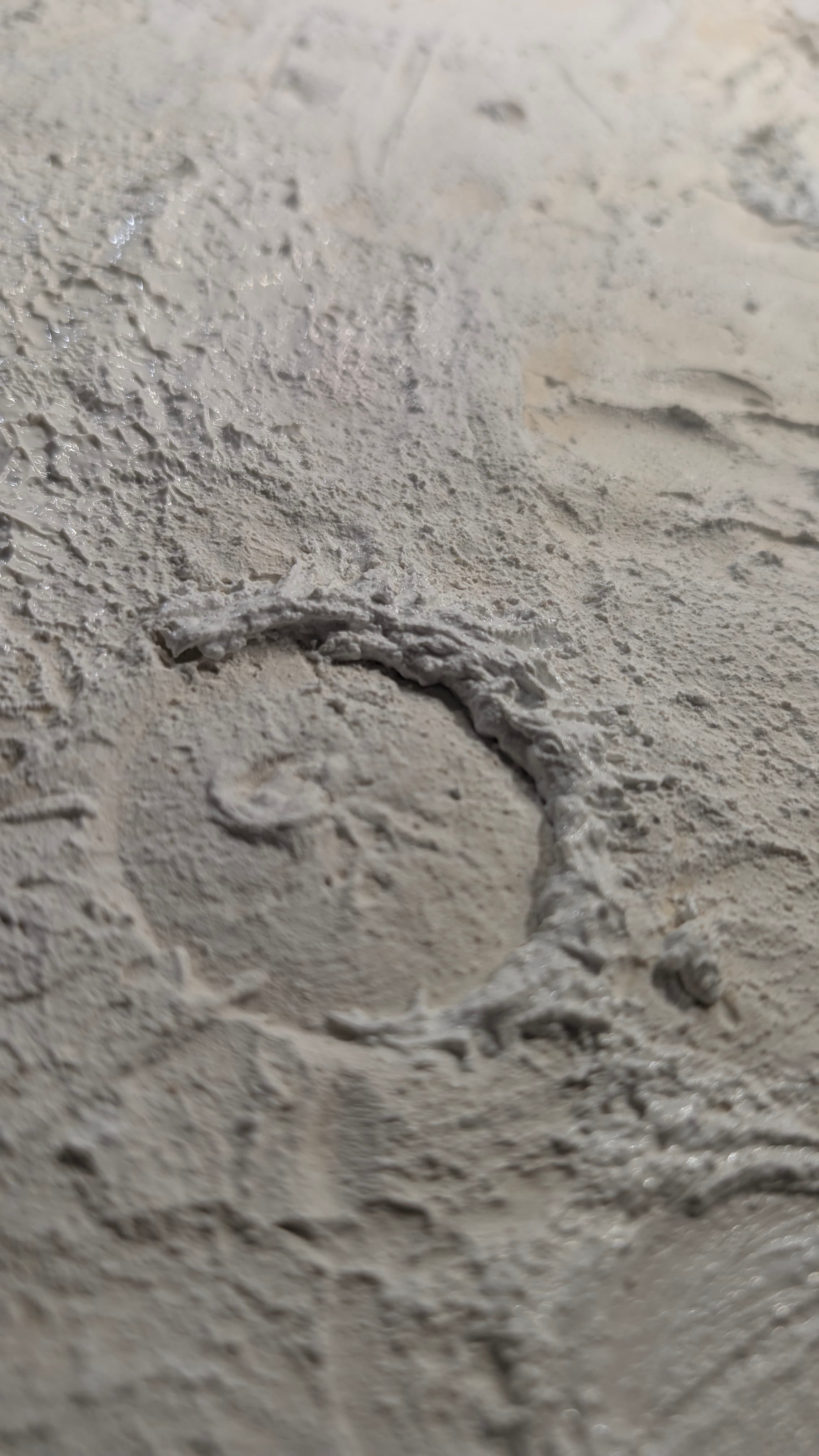 Weiße, grob strukturierte Oberfläche mit einer kreisförmigen, kraterähnlichen Erhebung in der Bildmitte.