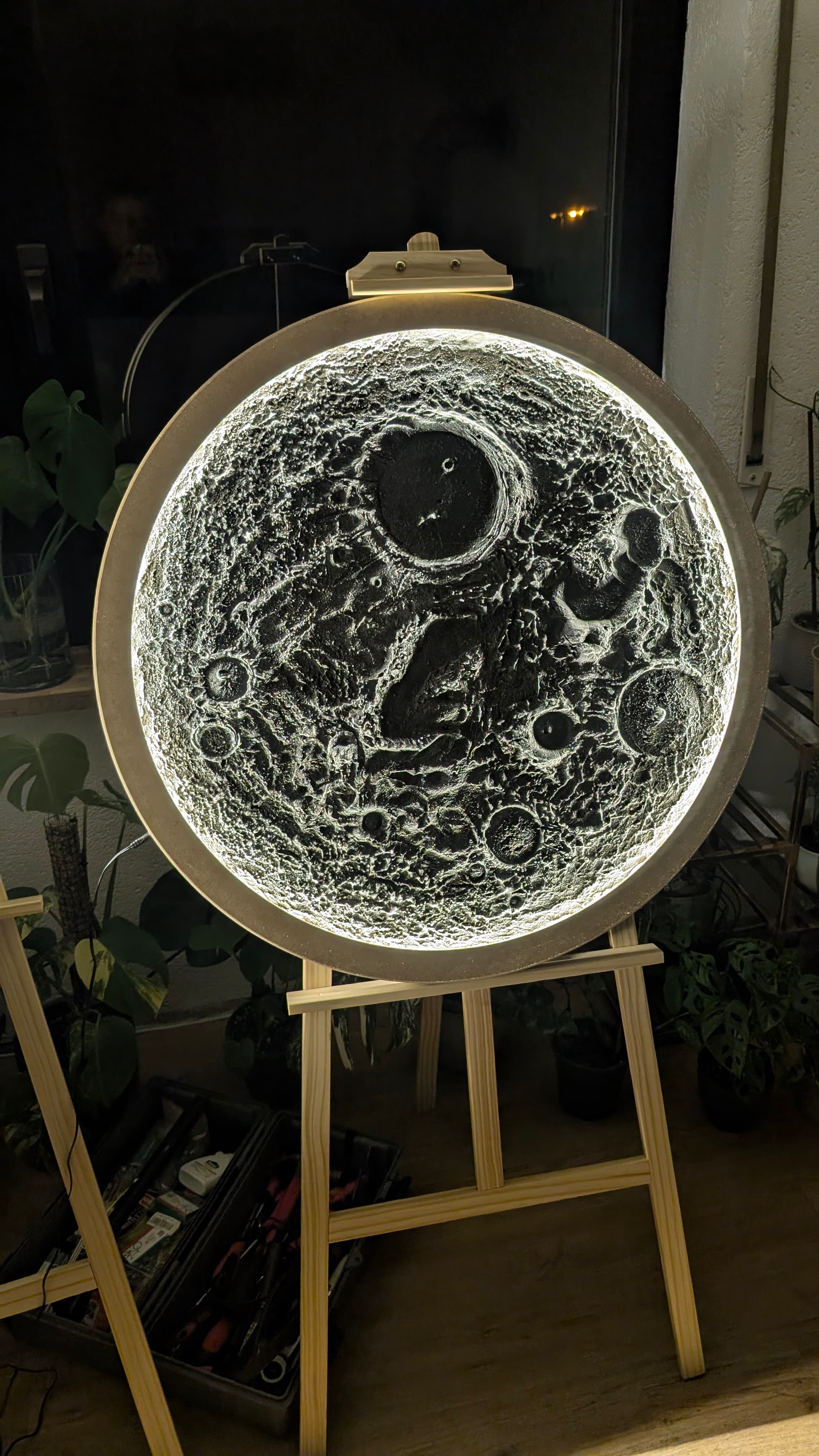 Eine leuchtende, kreisrunde Mondoberfläche mit Kratern auf einer hölzernen Staffelei in dunkler Umgebung.