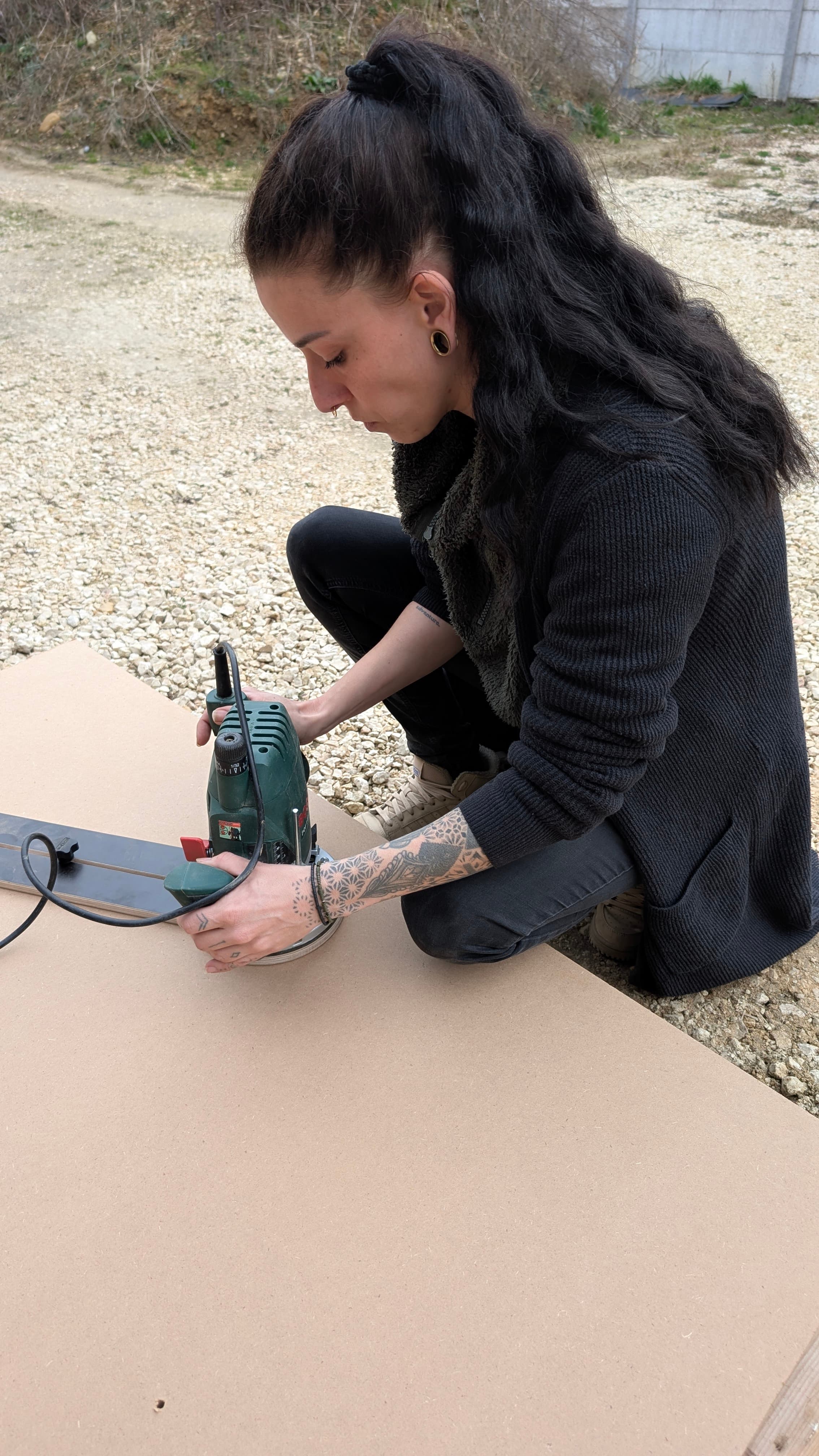 Tätowierte Frau mit langen dunklen Haaren bearbeitet eine Holzplatte im Freien mit einer grünen Oberfräse.