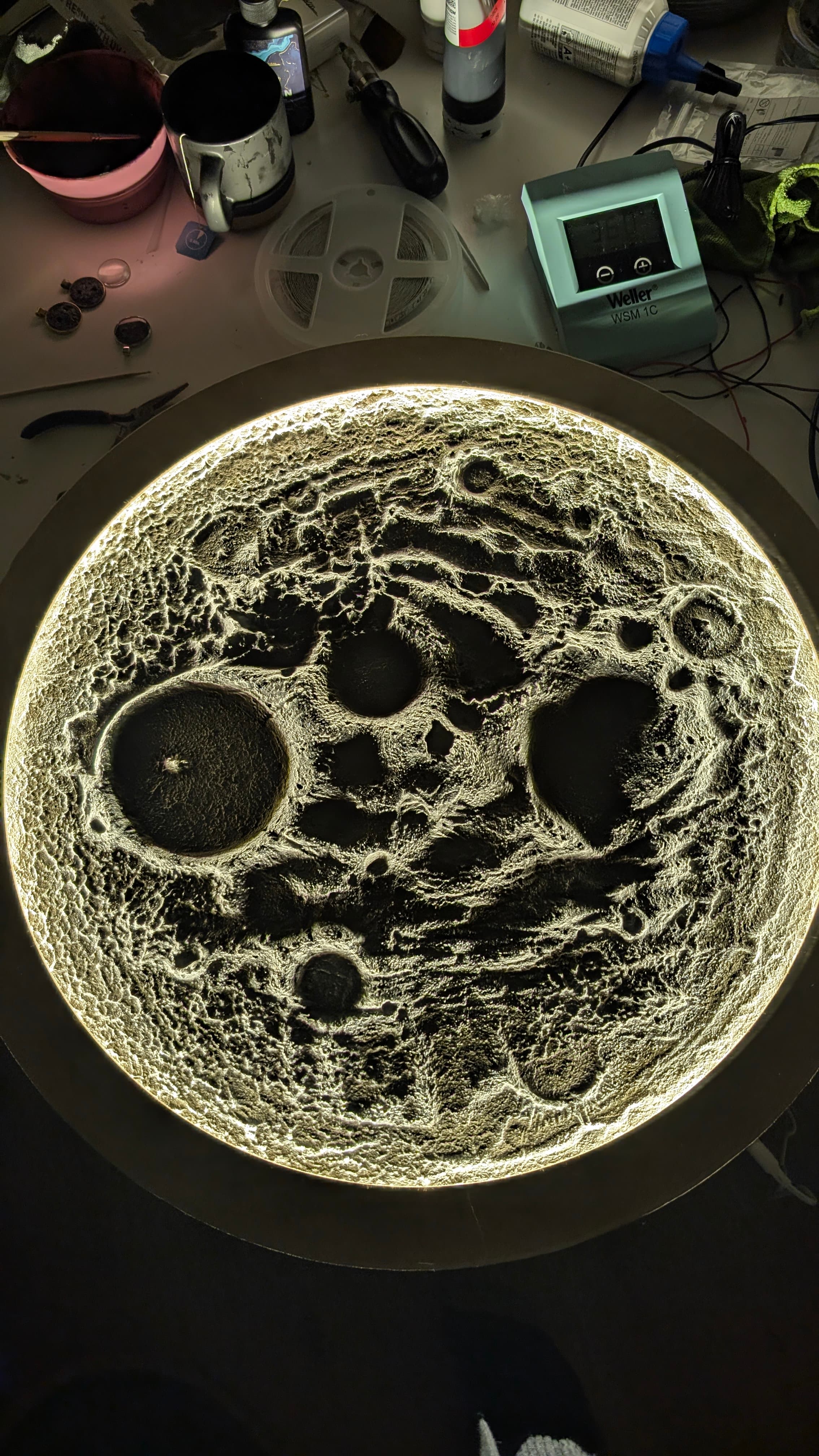 Hinterleuchtete, detailreiche Mondoberfläche mit Kratern auf einem Arbeitstisch, umgeben von Werkzeugen und Bastelmaterialien.