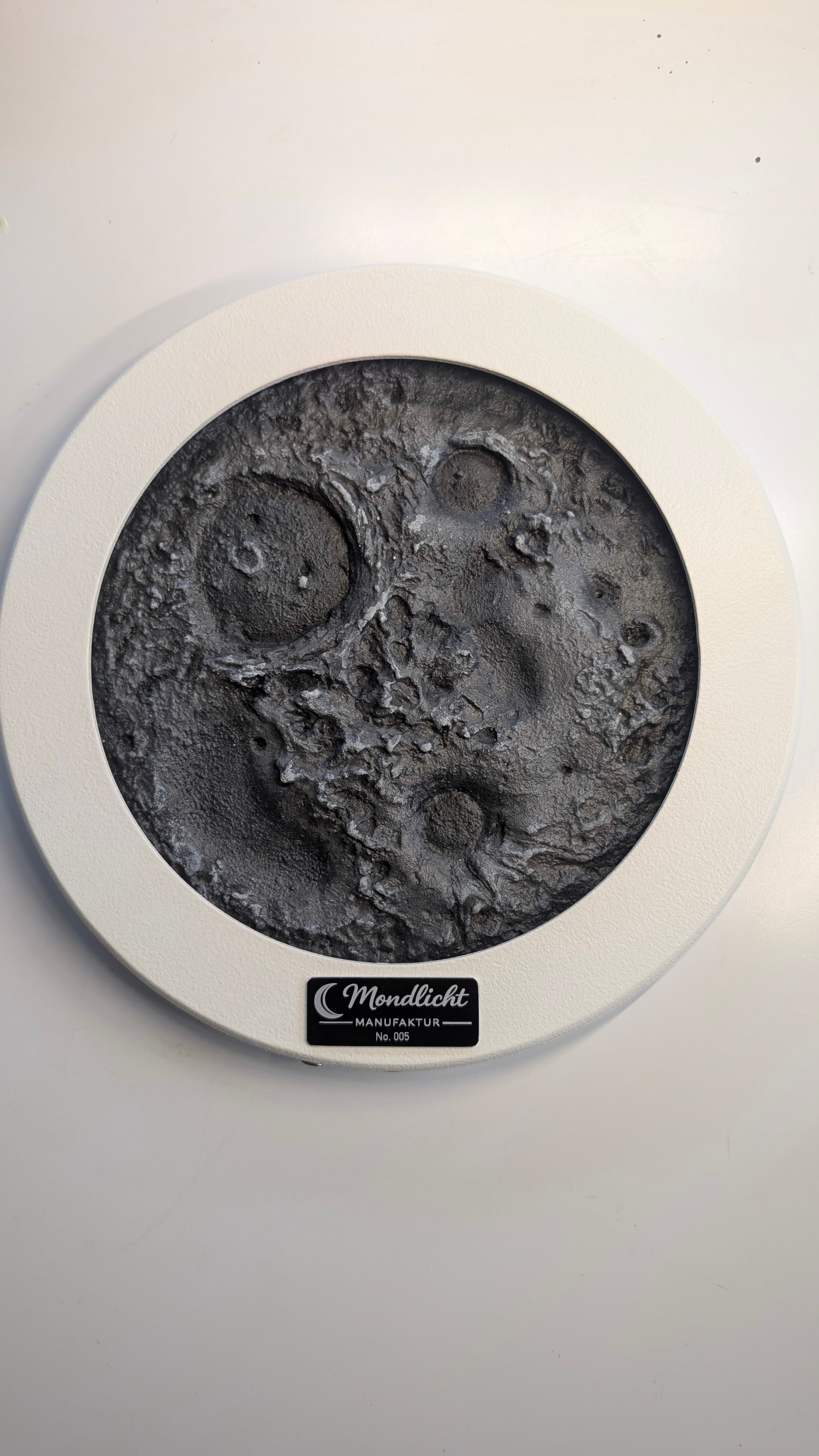 Detailreiche, graue Mondoberfläche mit Kratern, eingefasst in einen runden weißen Rahmen mit Herstellerplakette.