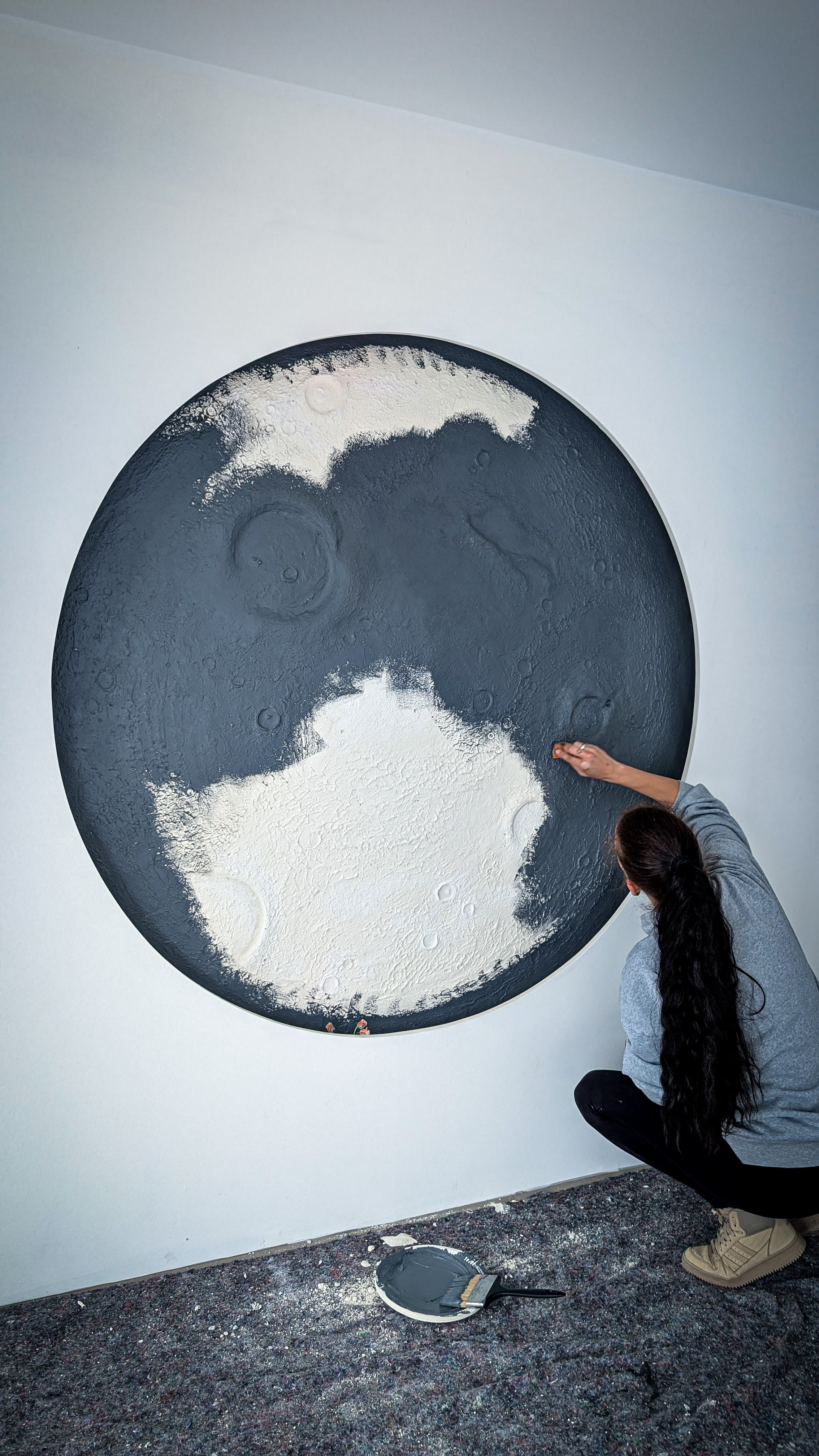 Frau gestaltet ein großes, texturiertes Mond-Wandbild in Grau und Weiß auf einer weißen Wand.