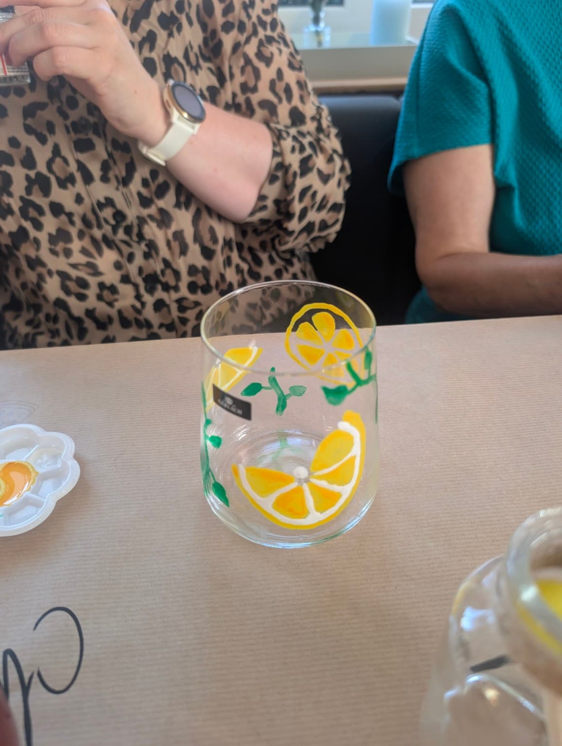 Ein handbemaltes Glas mit gelben Zitronenscheiben und grünen Blättern auf einem hellen Tisch.
