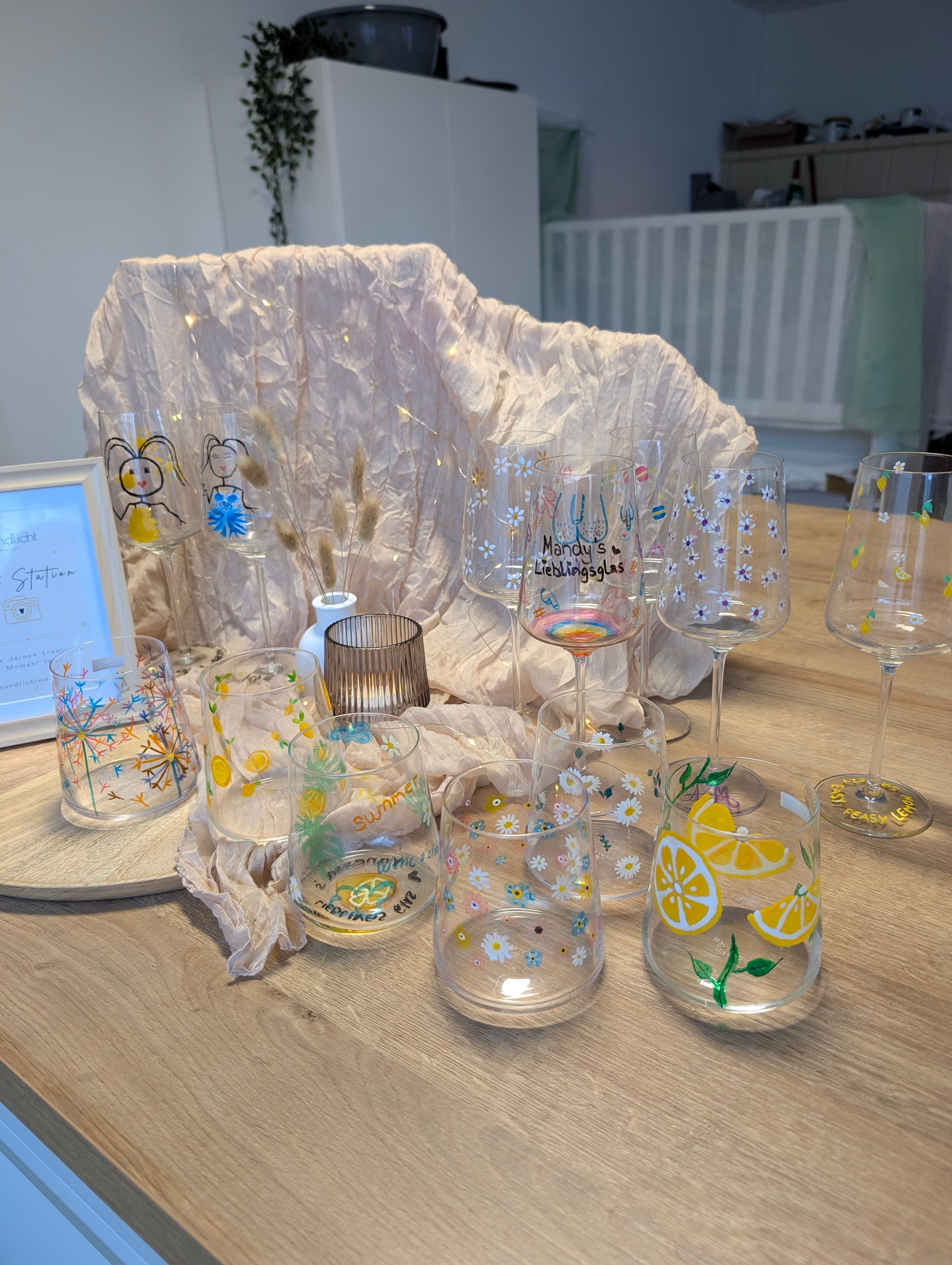 Handbemalte Weingläser und Becher mit bunten Blumen- und Zitronenmotiven auf einem hellen Holztisch.