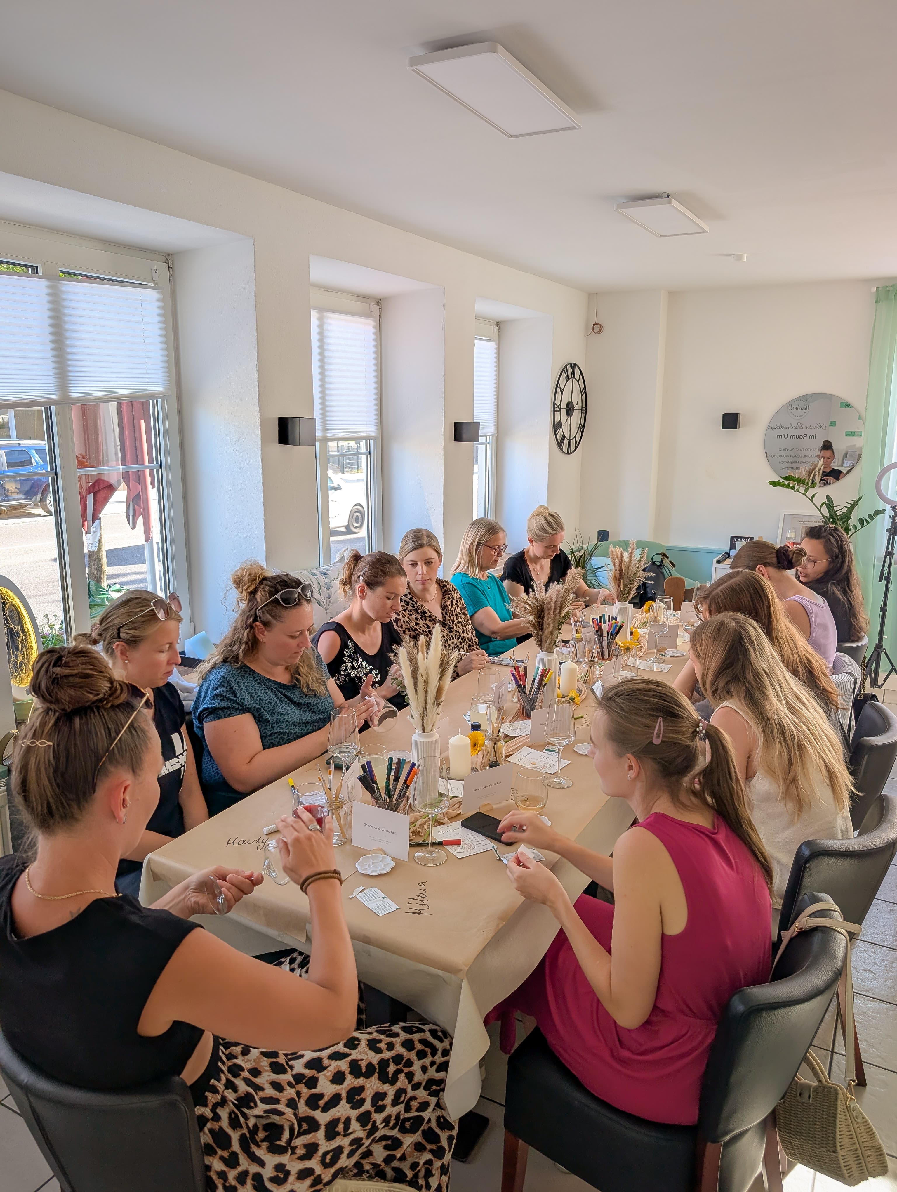 Frauen sitzen an einem langen Tisch und bemalen Weingläser bei einem Workshop im hellen Raum.