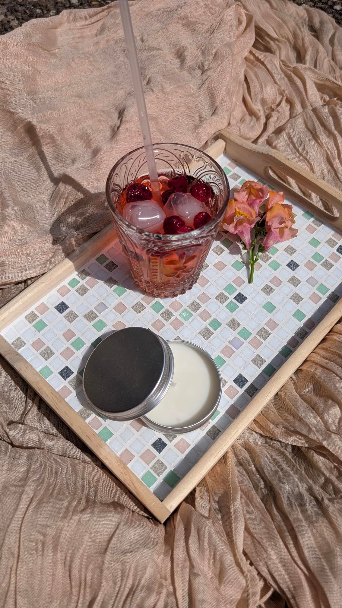 Mosaik-Tablett mit Beerengetränk, Eiswürfeln, offener Kerze und rosa Blume auf gerafftem beigem Stoff.
