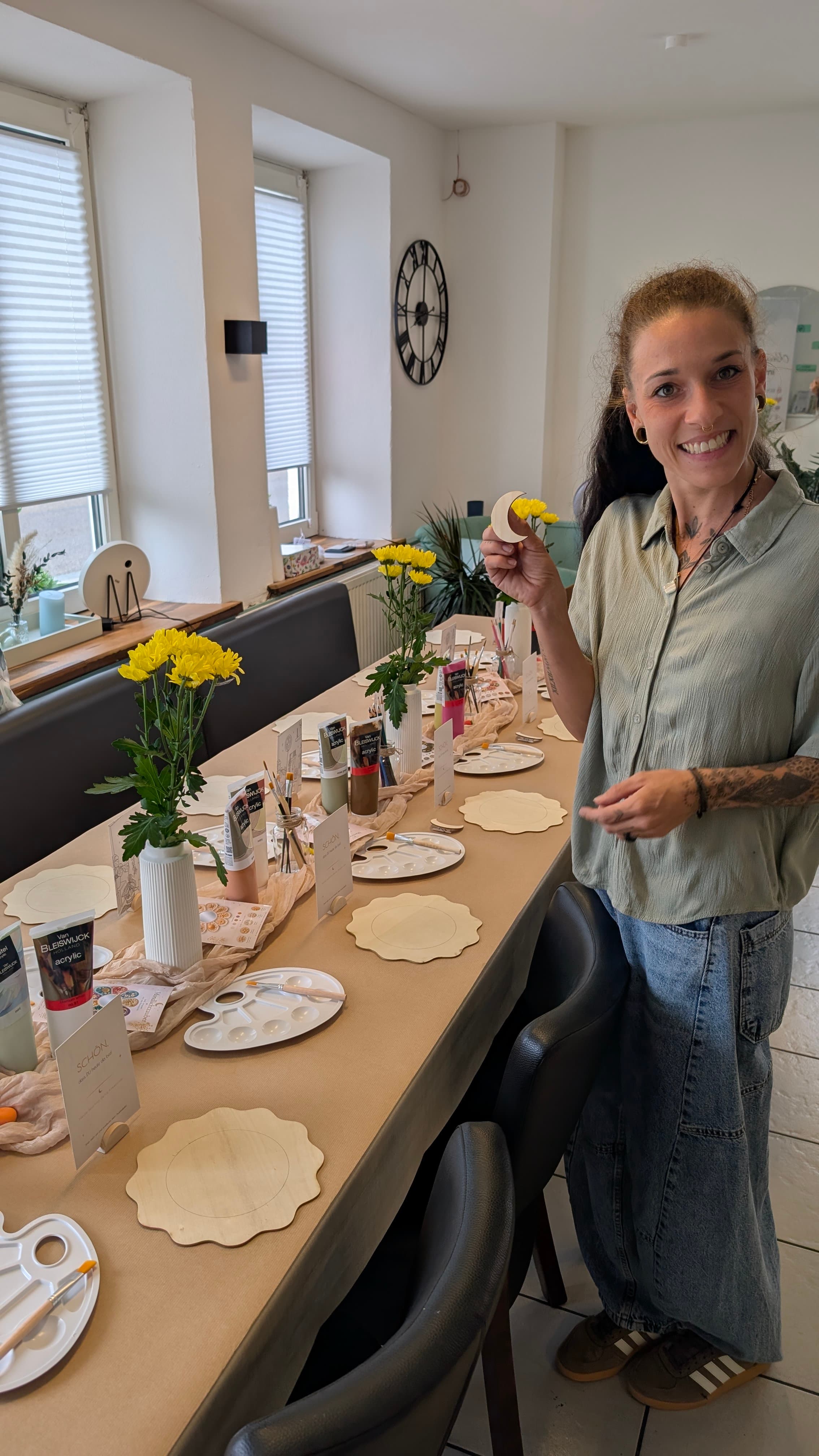 Lächelnde Frau hält eine Mondsichel an einem Tisch mit Malutensilien und gelben Blumen.