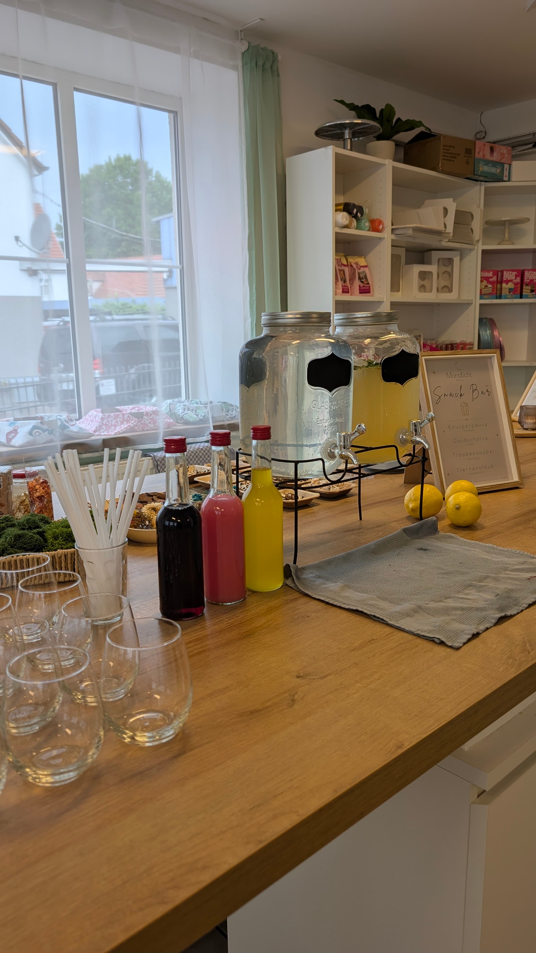 Getränkestation mit zwei großen Glasspendern, bunten Sirupflaschen, Gläsern und Zitronen auf einer hellen Holztheke.