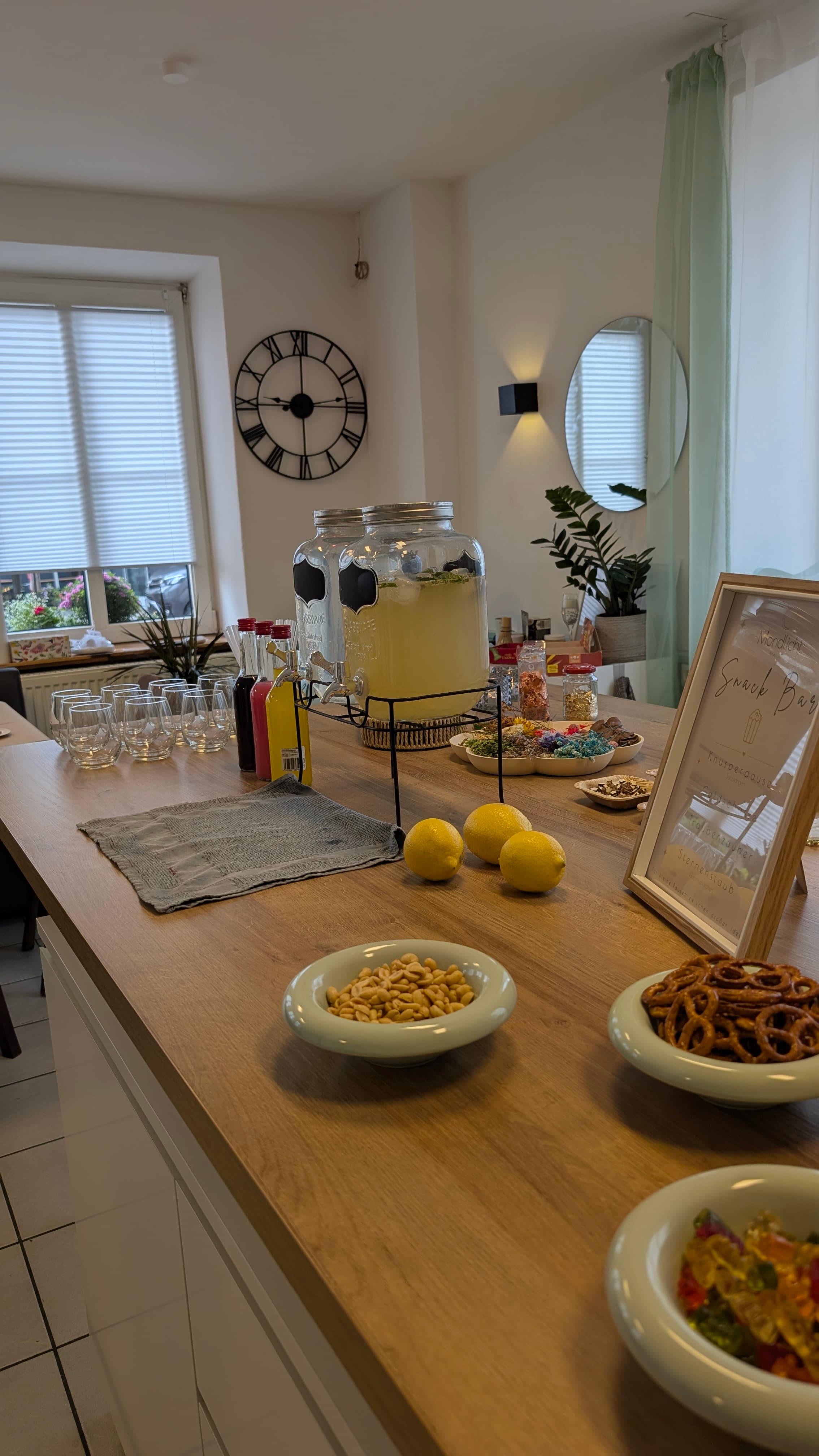 Snackbar auf Holztisch mit Getränkespendern, Gläsern, Zitronen und Schalen mit Erdnüssen, Brezeln und Gummibärchen.