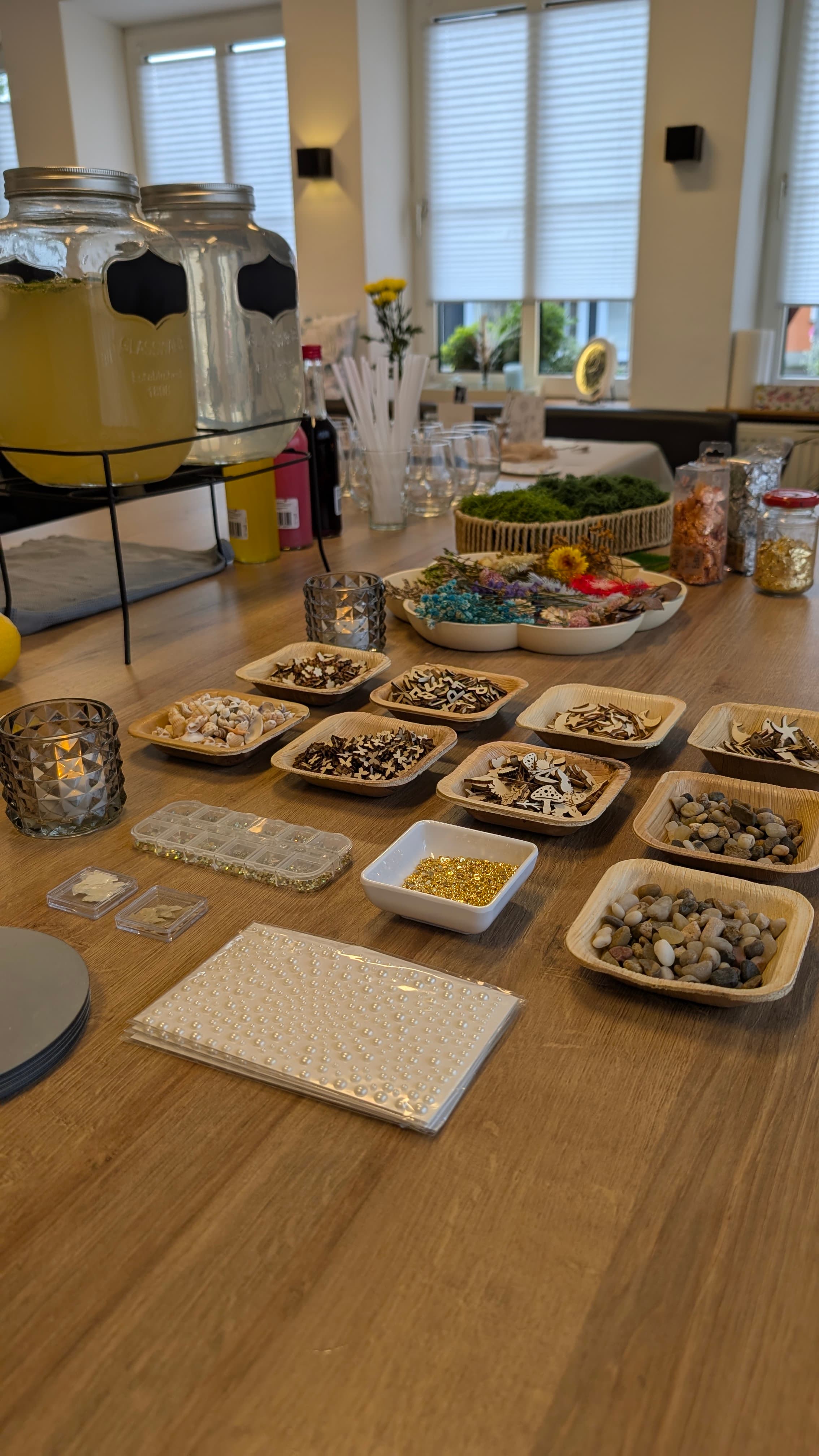 Holztisch mit kleinen Schalen voller Bastelmaterialien wie Muscheln, Steinen, Holzformen und goldenen Perlen.