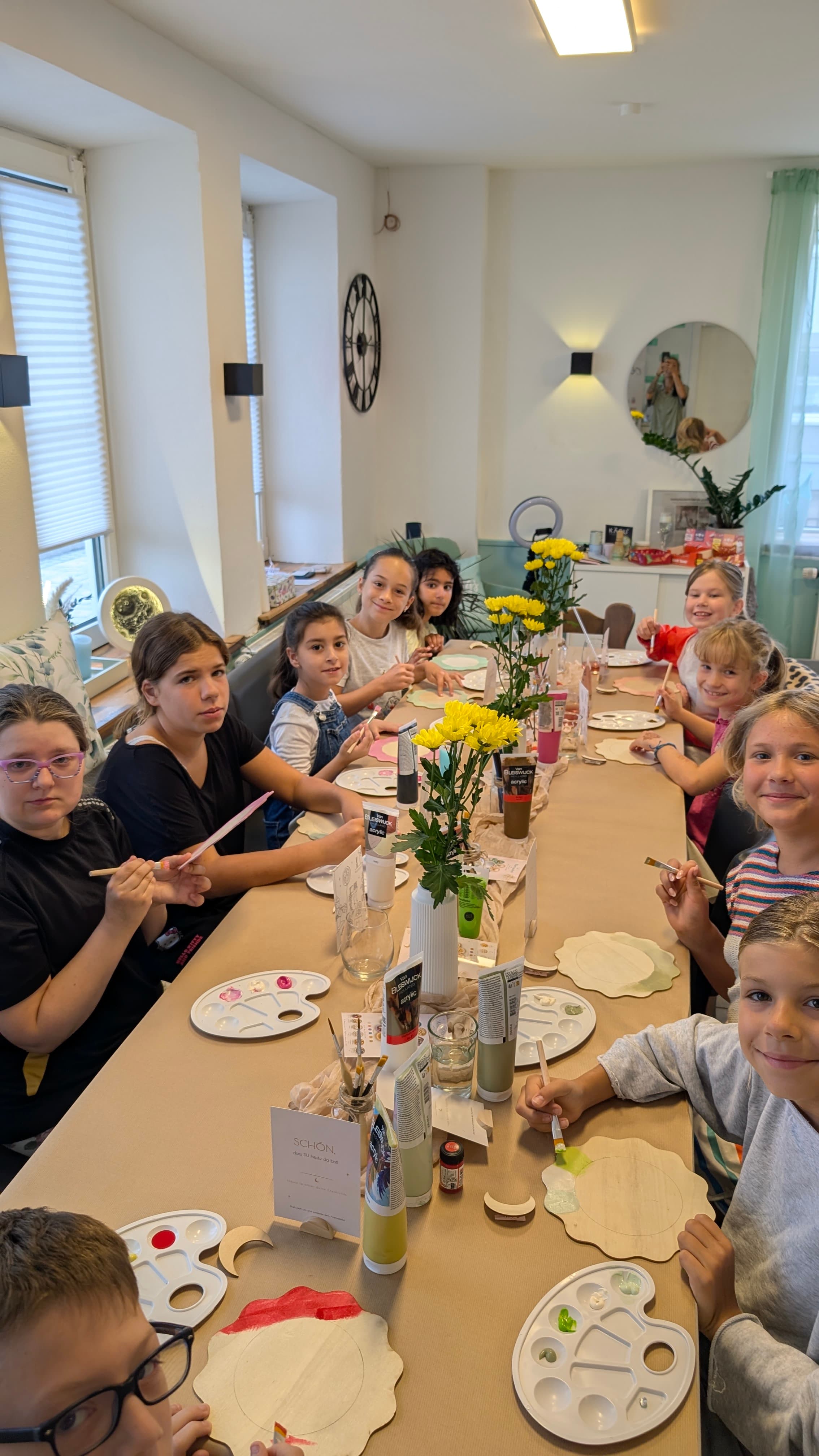 Kinder sitzen an einem langen Tisch und malen mit Pinseln auf verschiedene Holzformen.