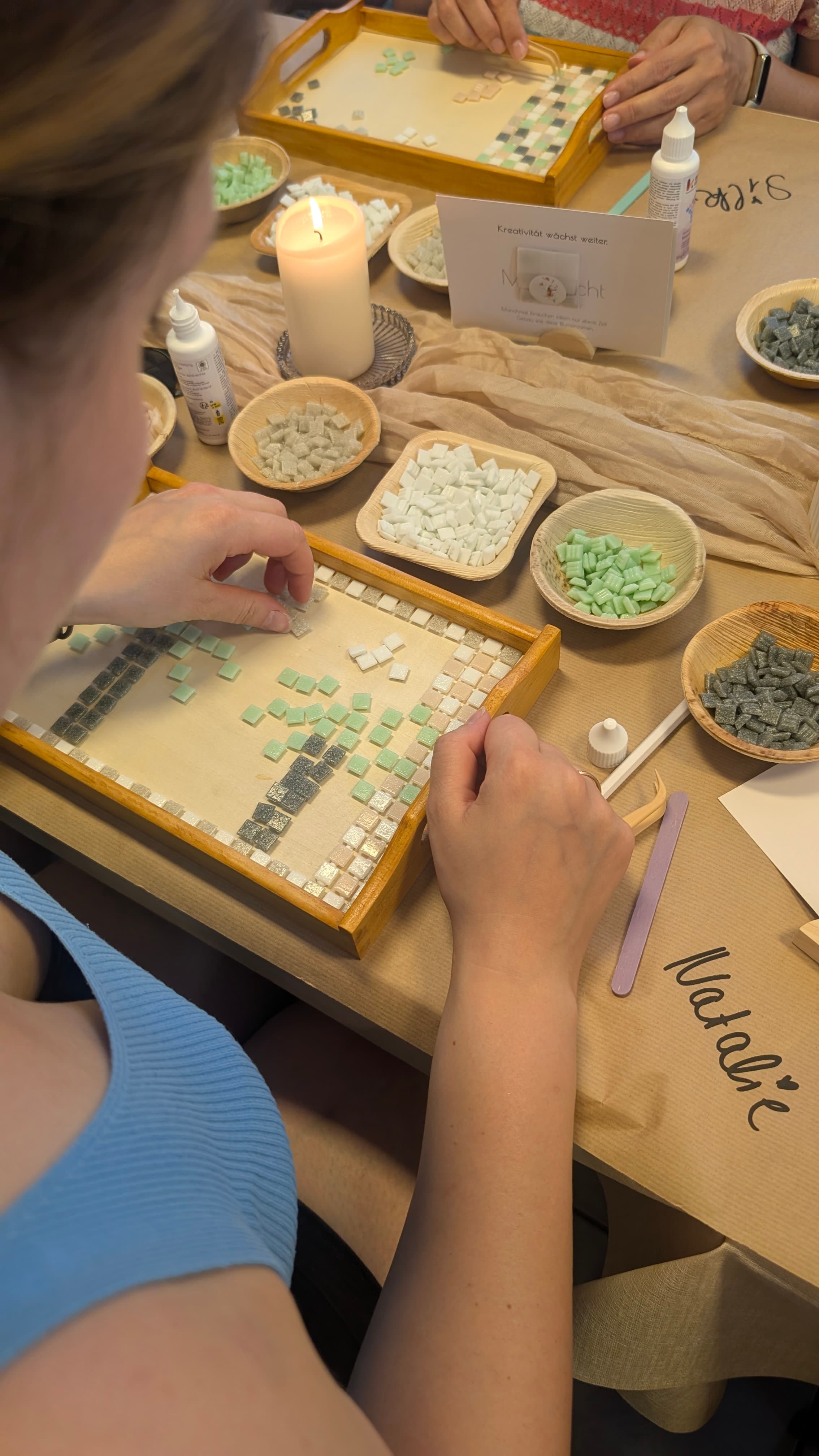 Eine Frau legt grüne, graue und weiße Mosaiksteine auf ein Holztablett neben einer brennenden Kerze.