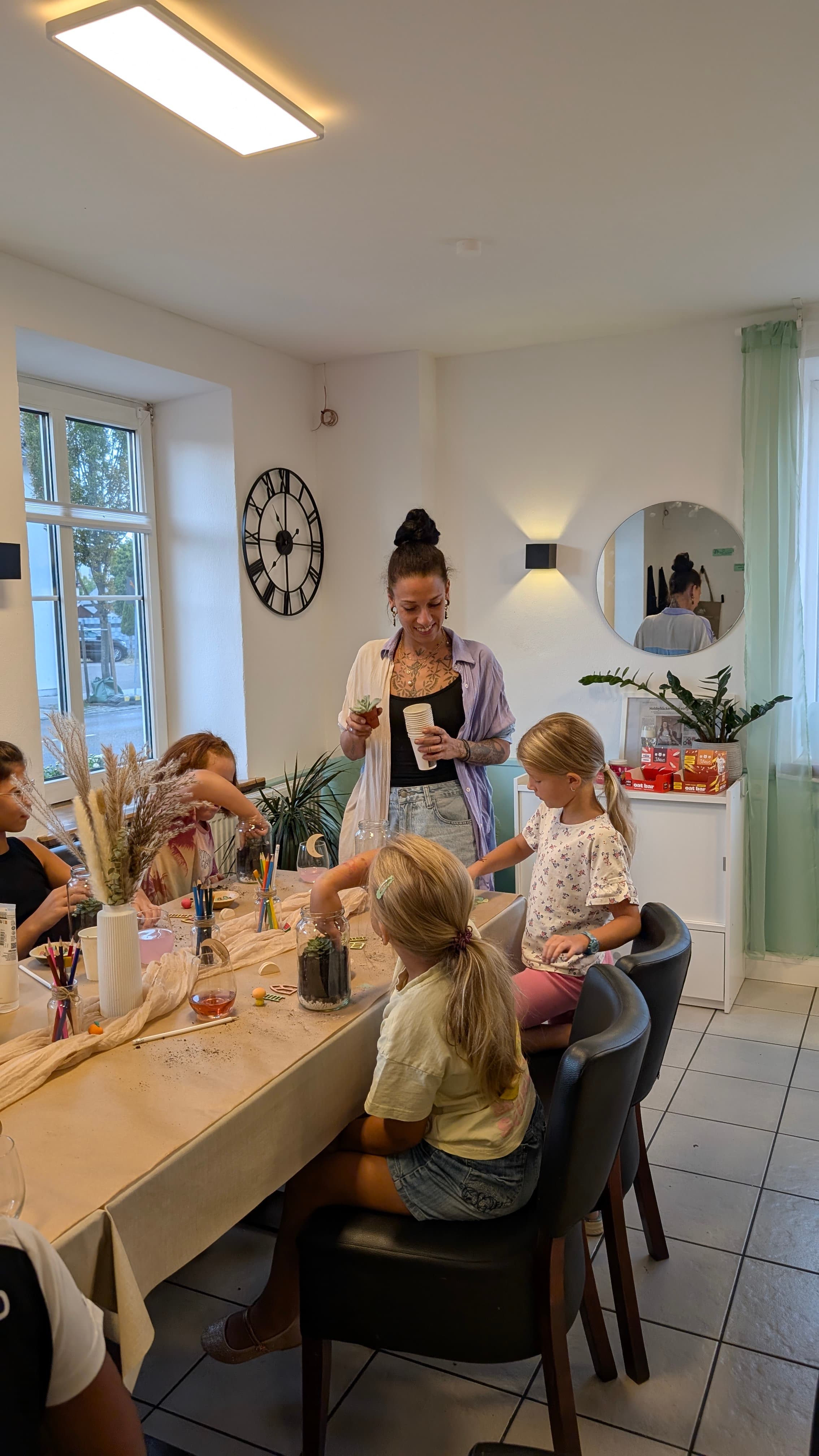 Eine Frau und Kinder basteln gemeinsam an einem Tisch kleine Terrarien mit verschiedenen Pflanzen.