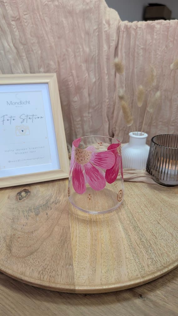 Handbemaltes Glas mit rosa Blütenblättern auf einem Holztablett neben einem Bilderrahmen und Trockenblumen.