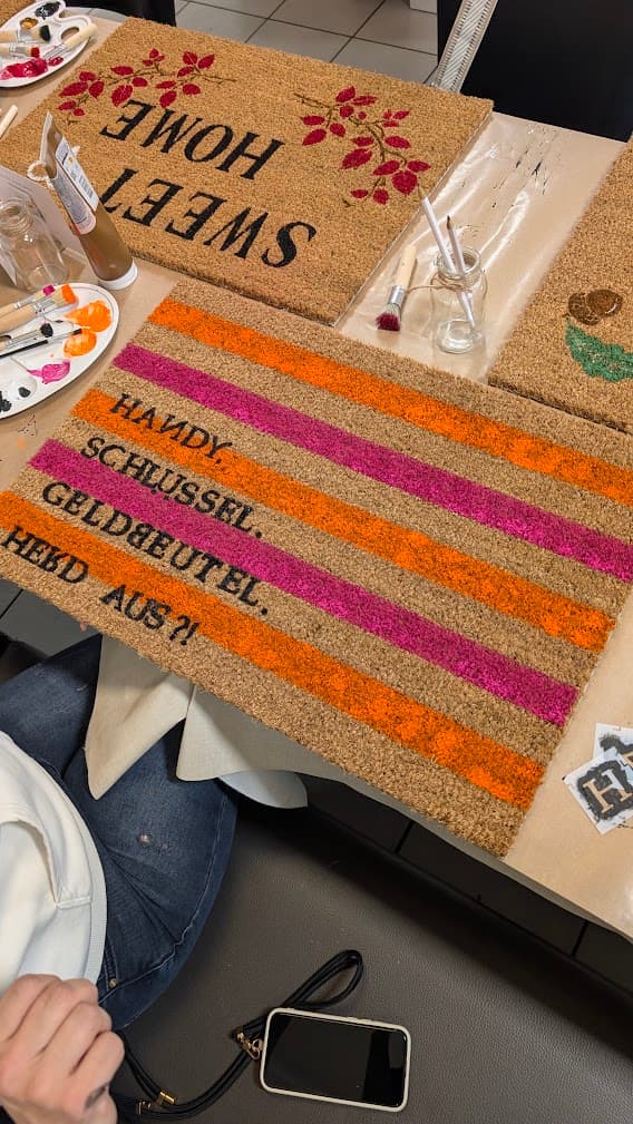 Handbemalte Kokosmatten mit Schriftzügen und bunten Streifen neben Pinseln und Farbpaletten auf einem Arbeitstisch.