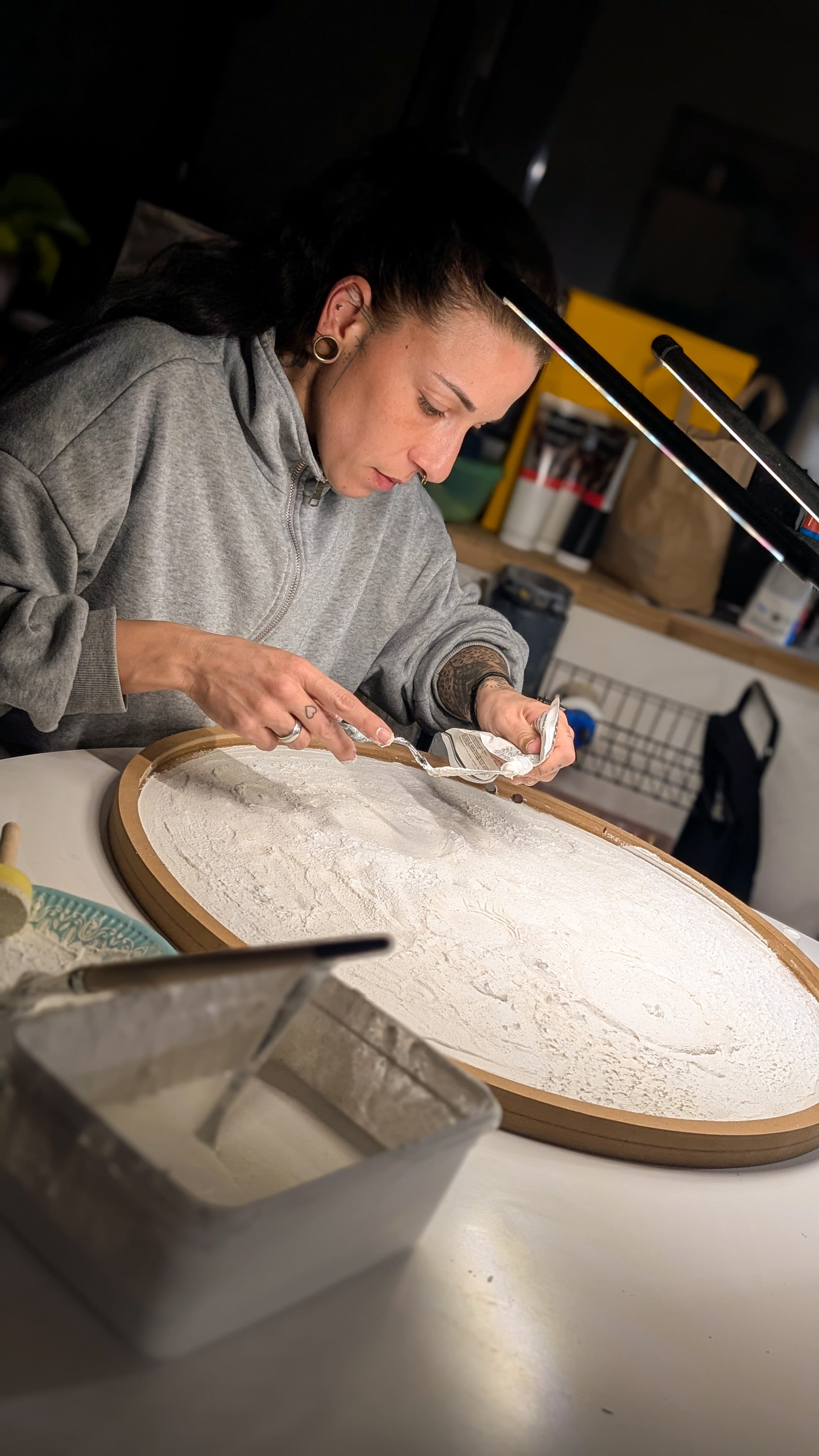 Künstlerin im grauen Hoodie trägt konzentriert weiße Strukturpaste auf ein rundes, hölzernes Kunstwerk auf.