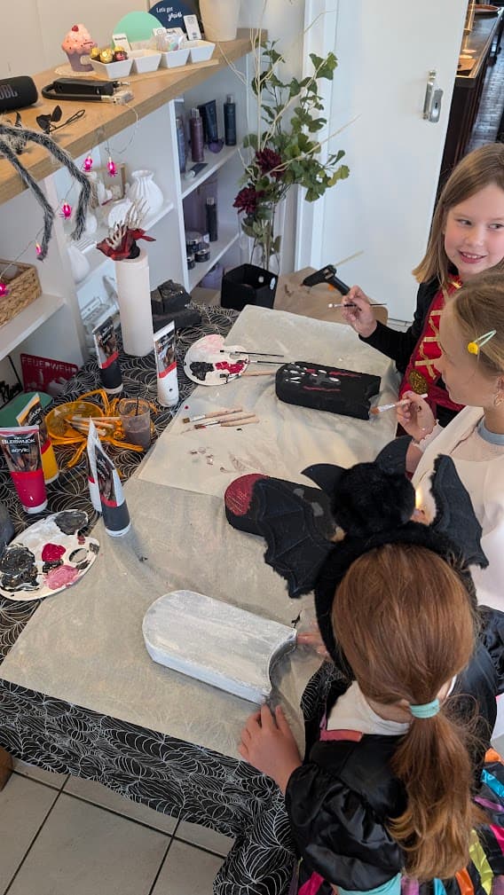 Drei Kinder bemalen an einem Tisch mit Spinnennetz-Tischdecke kleine Grabsteine mit Acrylfarben für Halloween.