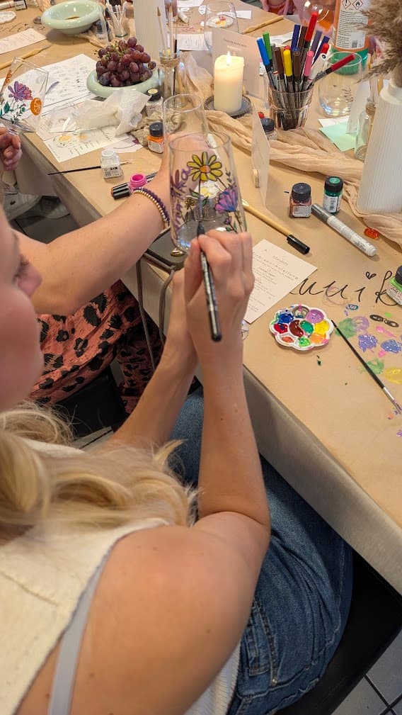 Blonde Frau bemalt ein Glas mit bunten Blumenmotiven an einem Tisch voller kreativer Malutensilien.