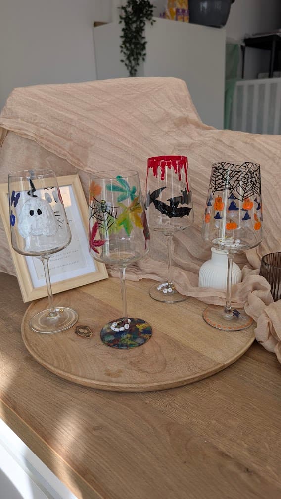Vier handbemalte Weingläser mit Halloween-Motiven wie Geistern, Fledermäusen und Kürbissen auf einem Holztablett.