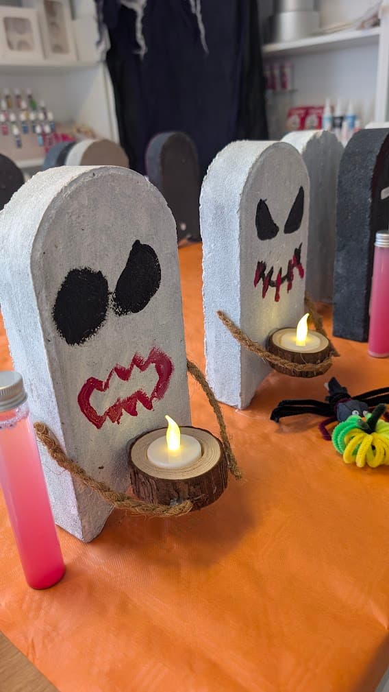 Zwei weiße Geisterfiguren mit gruseligen Gesichtern halten brennende LED-Teelichter auf einer orangefarbenen Tischdecke.