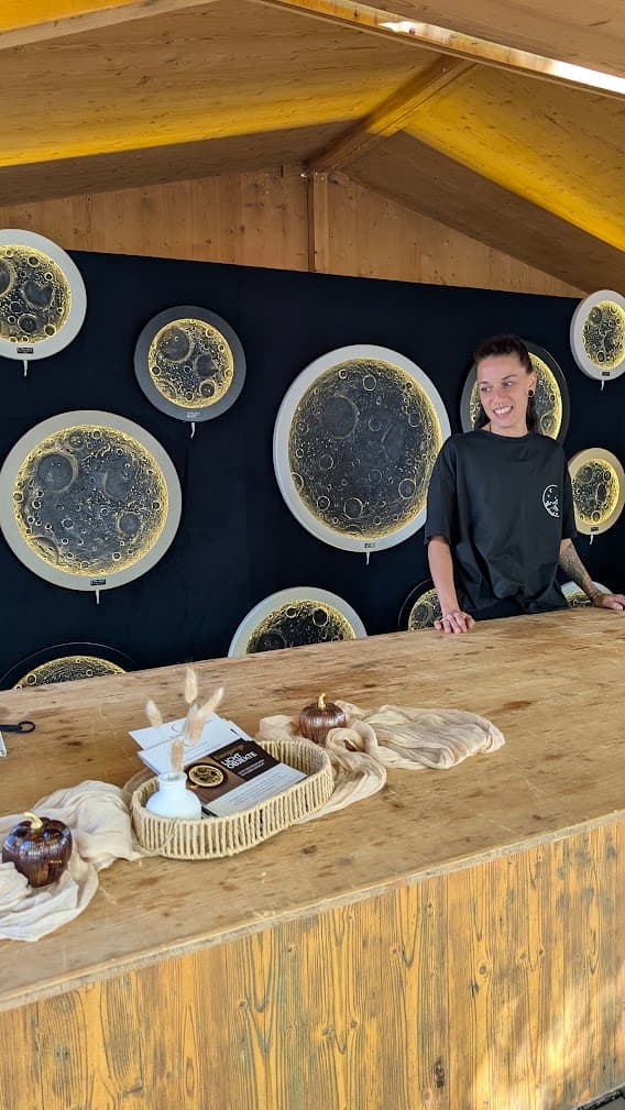 Lächelnde Frau hinter einem Holzstand präsentiert kunstvolle, beleuchtete Mond-Wandbilder auf schwarzem Hintergrund.