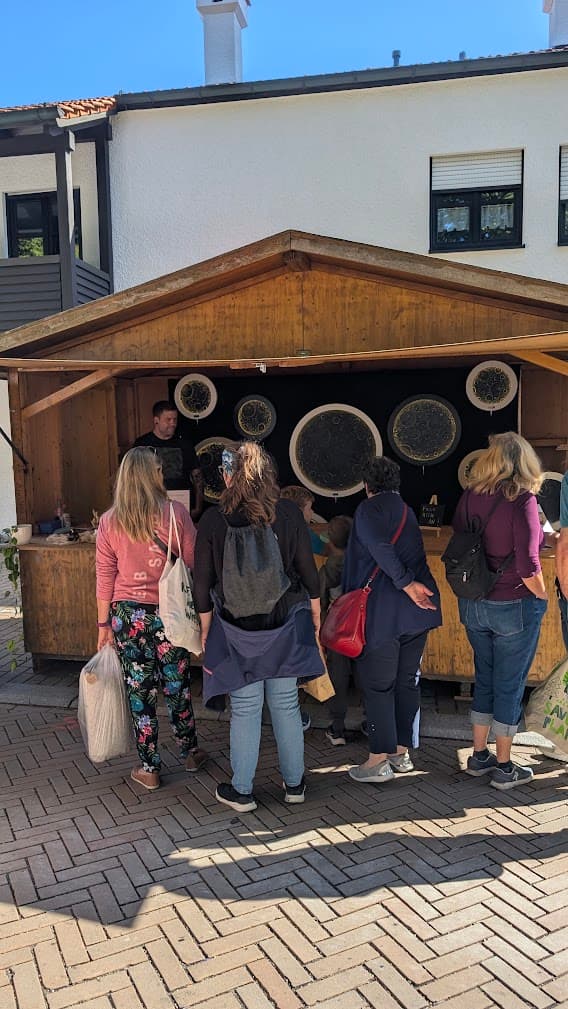 Menschen betrachten kreisförmige Kunstwerke an einem hölzernen Marktstand vor einem weißen Gebäude bei Sonnenschein.
