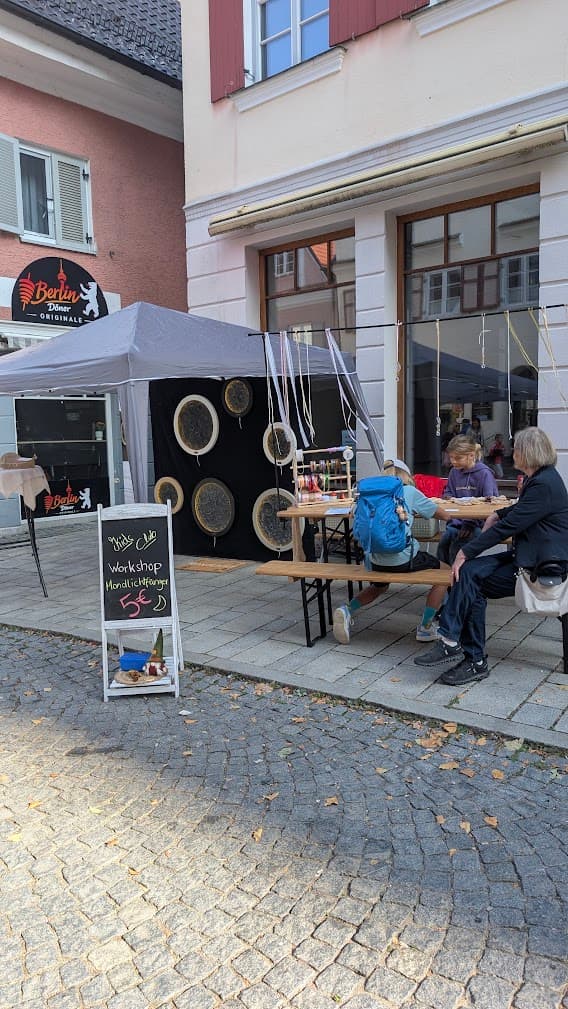 Kinder basteln Mondlichtfänger an einem Tisch unter einem grauen Pavillon in einer gepflasterten Straße.