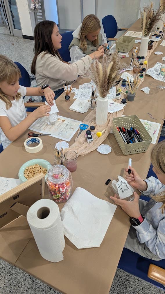Mädchen bemalen Gläser an einem langen Basteltisch mit Stiften, Farben und dekorativen Trockenblumen.