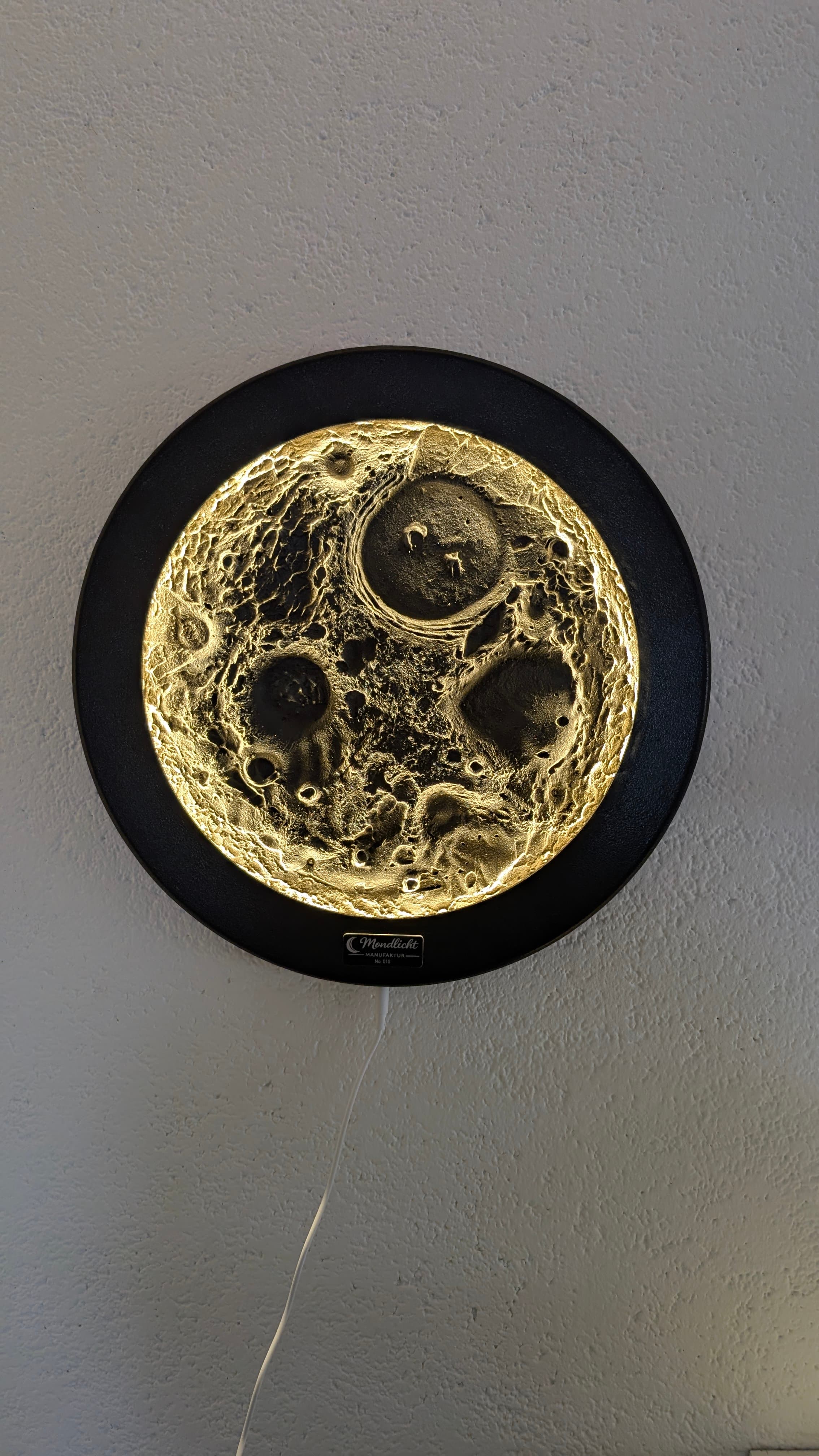 Kreisförmige, gold leuchtende Lampe mit strukturierter Mondoberfläche und schwarzem Rahmen an einer weißen Wand.