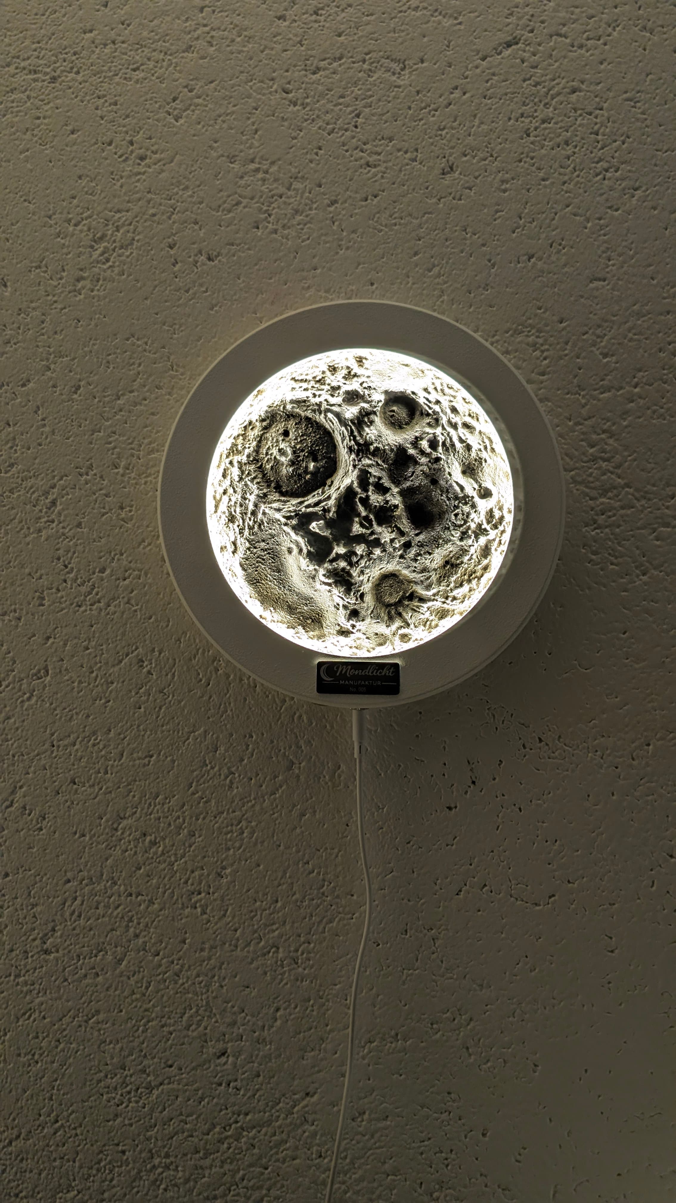 Eine hell leuchtende Wandlampe in Mondoptik mit detaillierten Kratern auf einer strukturierten, beigen Wand.