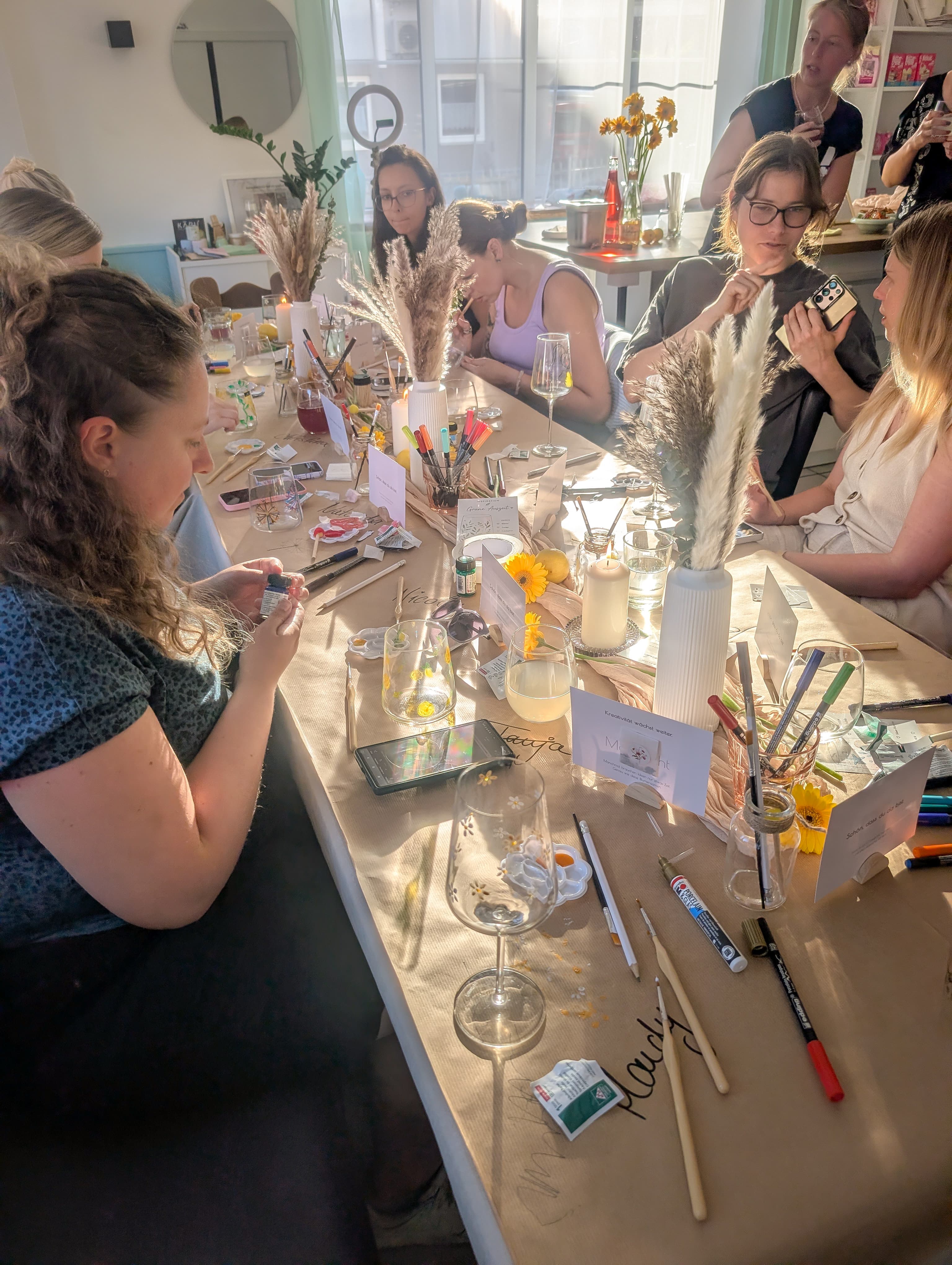 Frauen bemalen Gläser an einem langen Tisch mit Pinseln und Farben in hellem Sonnenlicht.