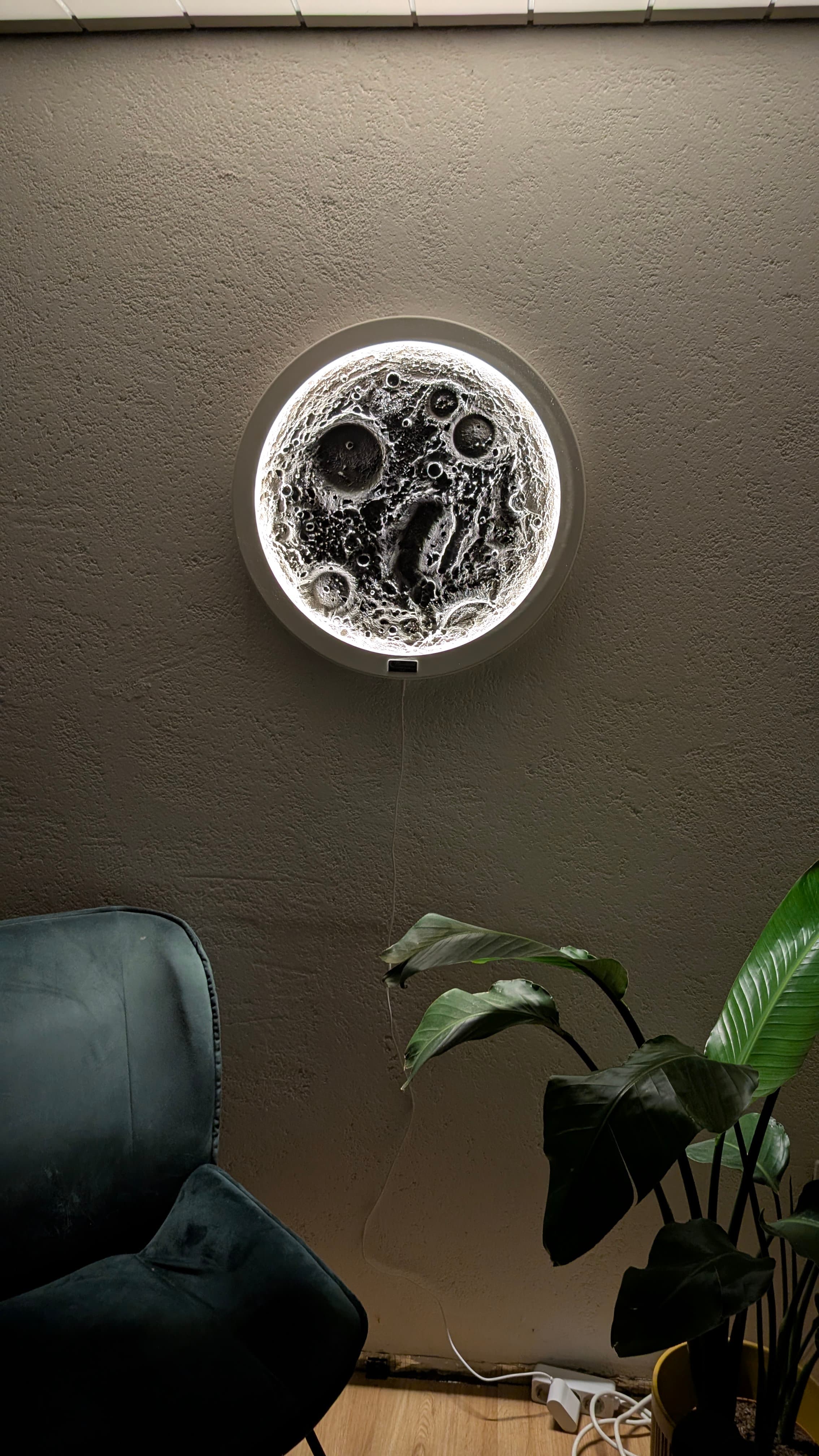 Eine leuchtende, kreisförmige Wandlampe in Mondoptik über einem grünen Sessel und einer großen Zimmerpflanze.