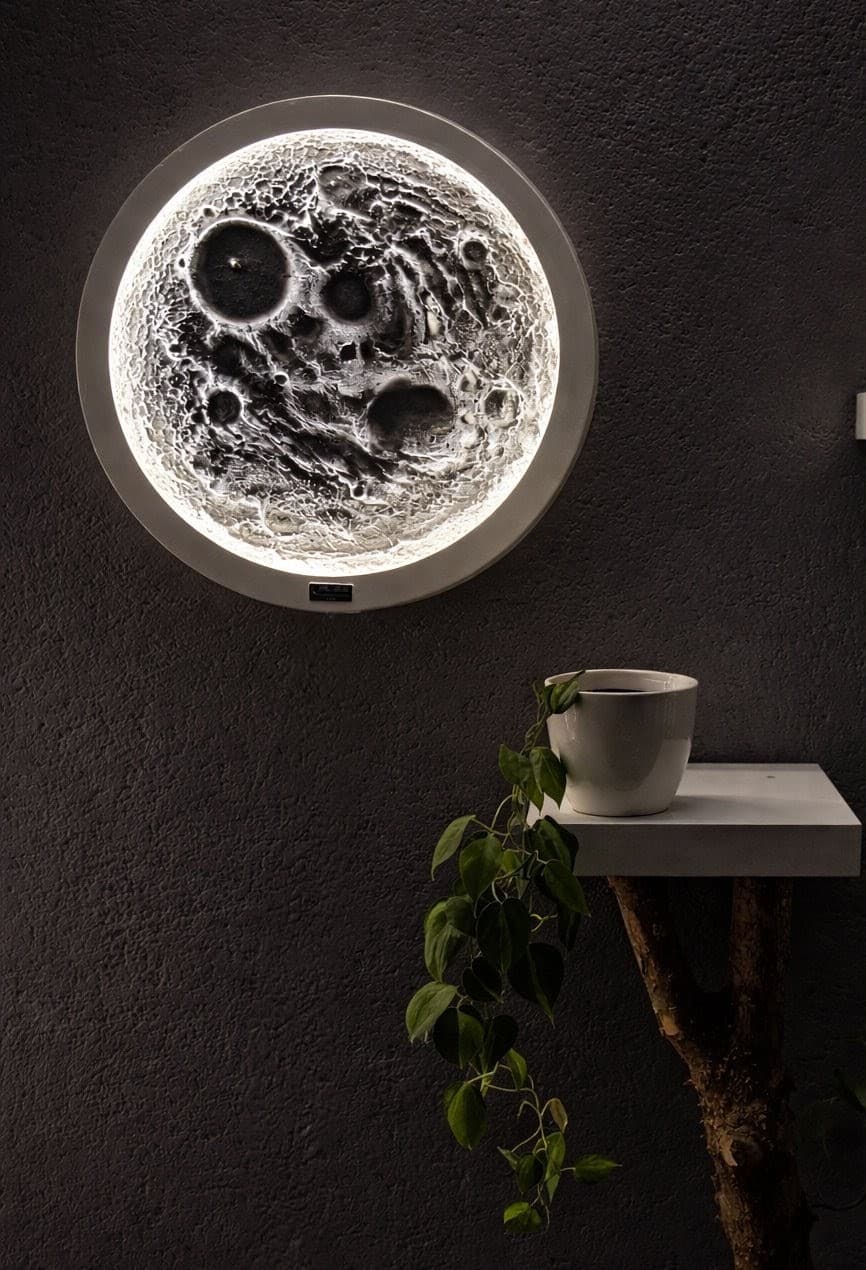 Runde, leuchtende Wandlampe in Mondoptik neben einer hängenden Pflanze auf einem kleinen weißen Regal.