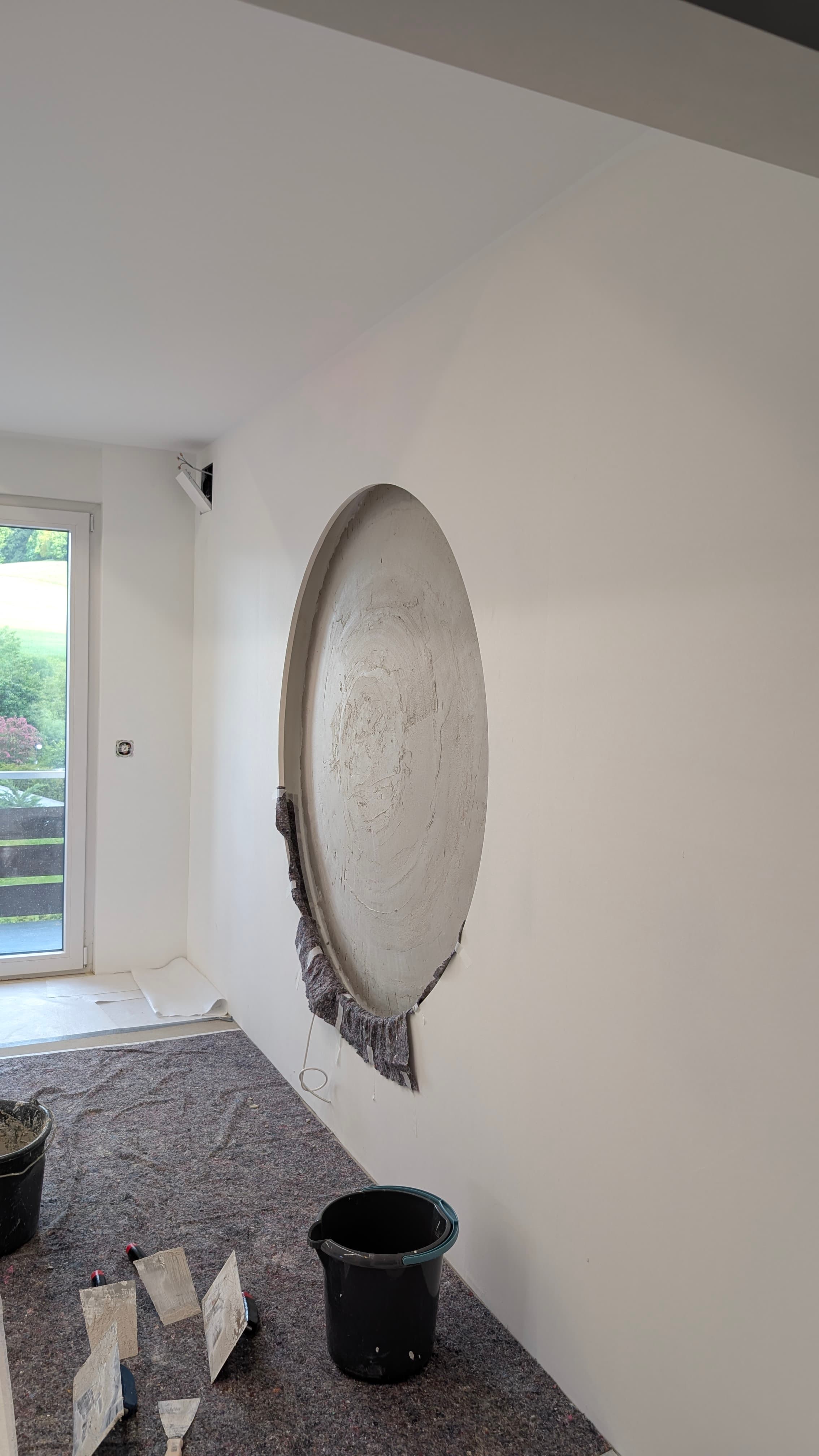 Weiße Wand mit kreisrundem Ausschnitt, frisch verputzt, daneben Eimer und Spachtel auf grauem Malervlies.