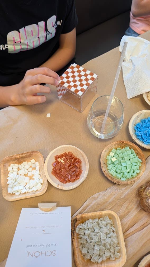 Eine Person beklebt einen Acrylwürfel mit Mosaiksteinen neben Schälchen mit bunten, quadratischen Steinchen.