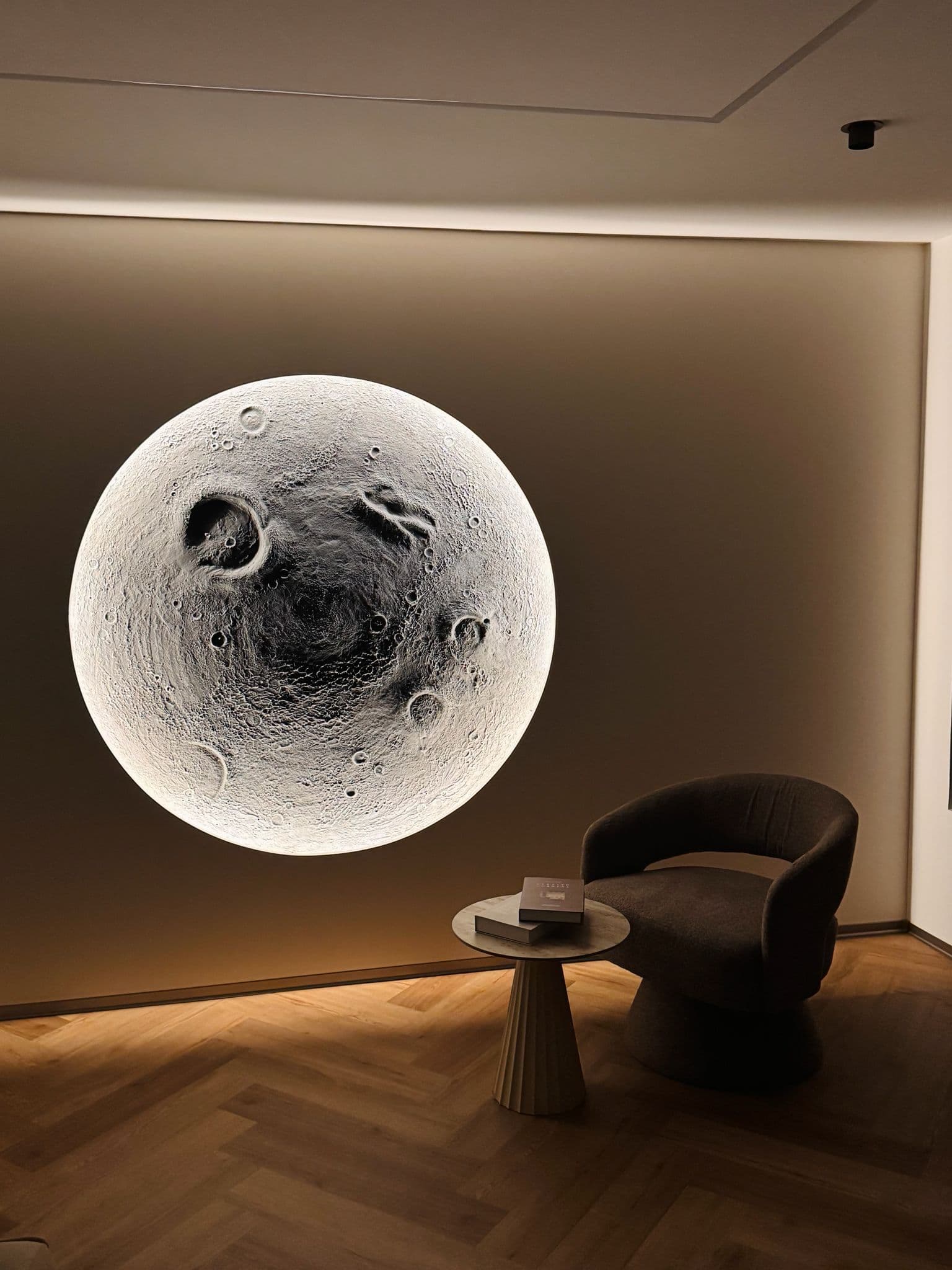 Große, leuchtende Mond-Wandlampe neben einem dunklen Sessel und Beistelltisch in einem modern gestalteten Raum.