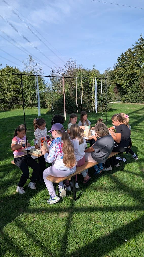 Kinder basteln an einem Holztisch im Freien vor einem Gestell mit hellen hängenden Bändern.