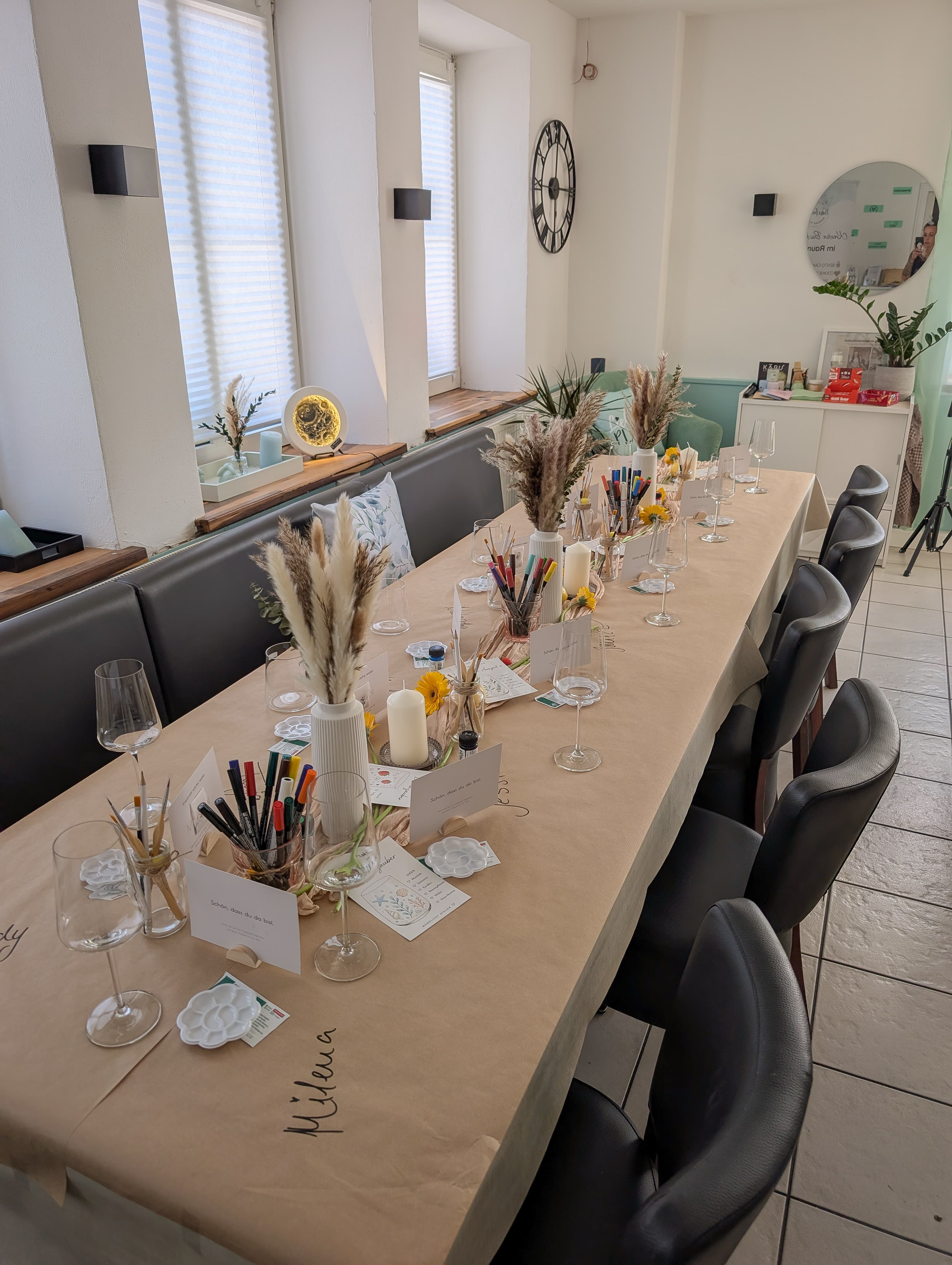 Gedeckter Tisch mit Malutensilien, Trockenblumen und Kerzen für einen kreativen Workshop in hellem Raum.